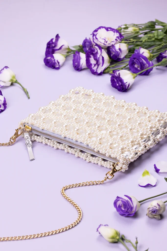 An elegant pearl clutch with a zipper closure and gold chain strap is styled with purple flowers for a soft, feminine aesthetic.