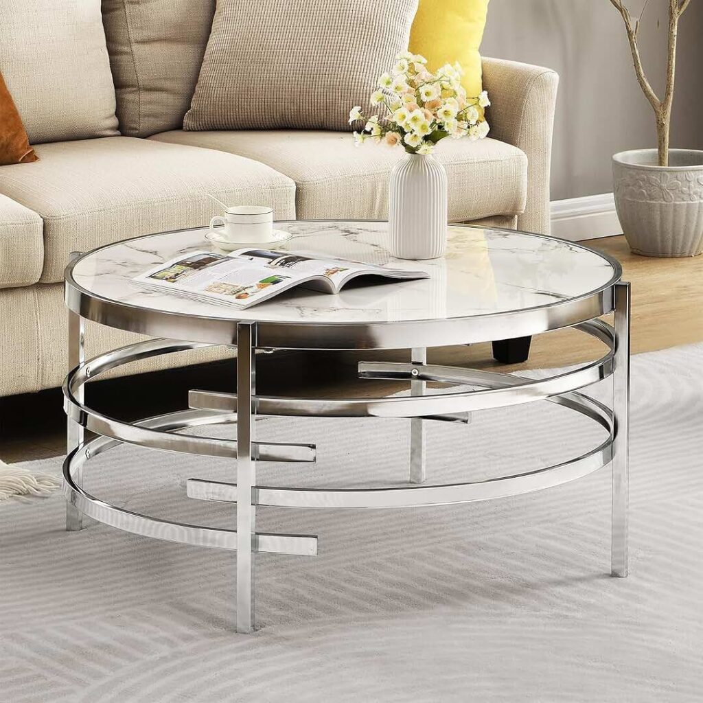 Round glass-top coffee table with a modern chrome metal frame, placed in front of a beige sofa. The table is decorated with a white vase of flowers, a teacup, and an open magazine.