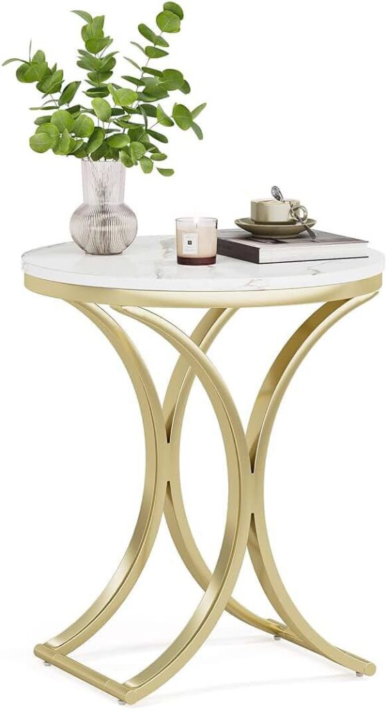 Elegant round side table with a white marble top and gold metal legs, decorated with a vase of green leaves, a scented candle, and a cup set on top of stacked books.