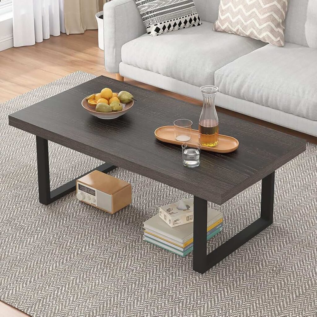 Modern rectangular wooden coffee table with black metal legs, featuring a bowl of fruit and a wooden tray with a glass carafe and cups. Books and a retro-style radio are stored on the lower shelf beneath the table.