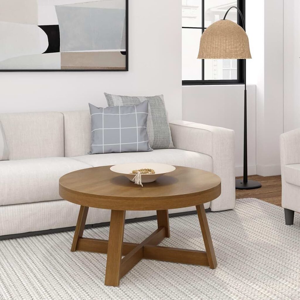 Round wooden coffee table with a minimalist design, placed in front of a light beige sofa. The table features a decorative bowl, and the room is accented with neutral tones, patterned throw pillows, and a woven floor lamp.