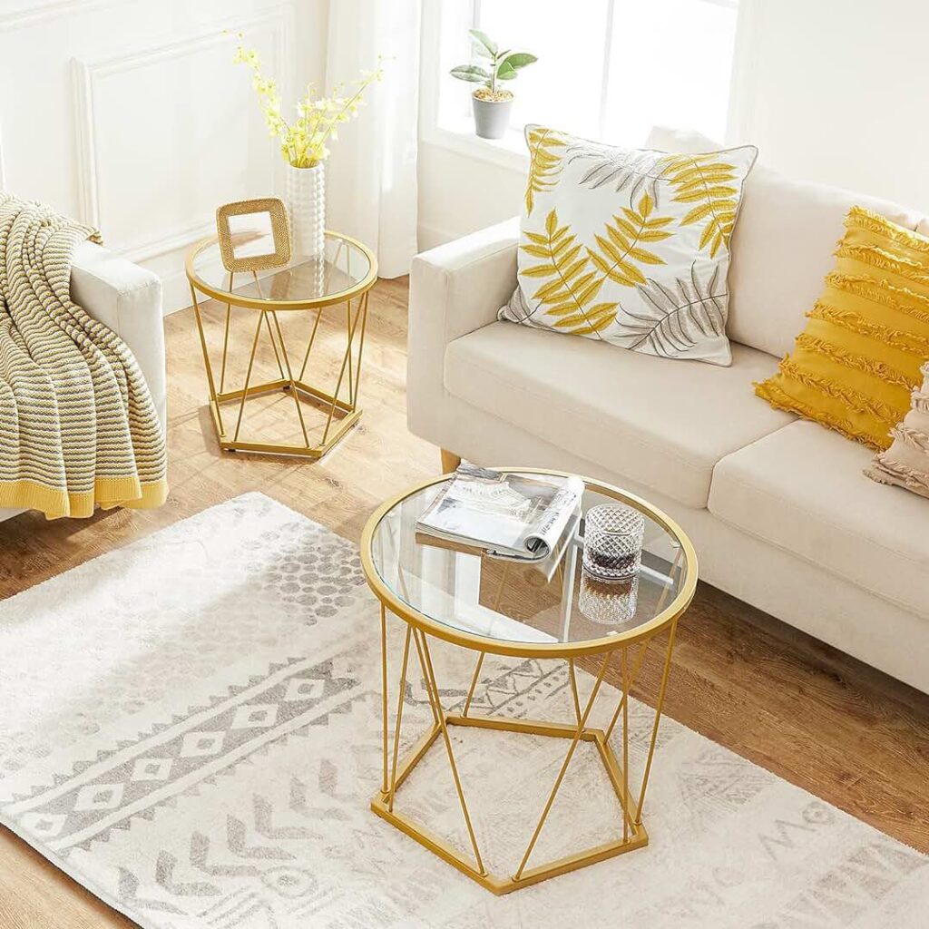 Bright living room with a beige sofa, decorated with yellow and white patterned pillows. Two gold-framed glass-top side tables are placed next to the sofa, one holding a magazine and candle, while the other displays a vase of yellow flowers and a decorative frame. The room features light-colored decor with a geometric-patterned rug.