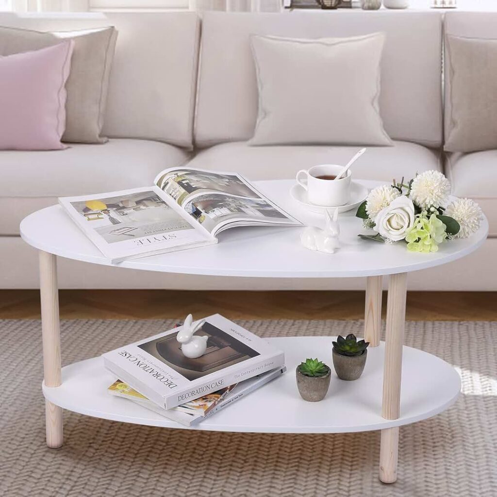 White oval coffee table with wooden legs, featuring a teacup, open magazine, and a flower arrangement. The lower shelf holds additional books and small succulent plants. The table is placed in front of a beige sofa with neutral and pink accent pillows.