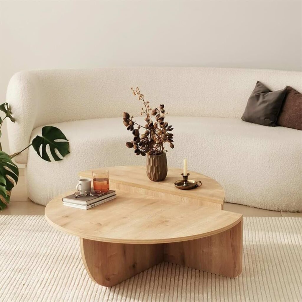 Contemporary living room featuring a light wooden coffee table with layered design, styled with a vase of dried flowers, a candle holder, and a cup of tea on books. The cozy space includes a white boucle sofa, dark accent pillows, and a soft cream rug, with a potted monstera plant in the corner.