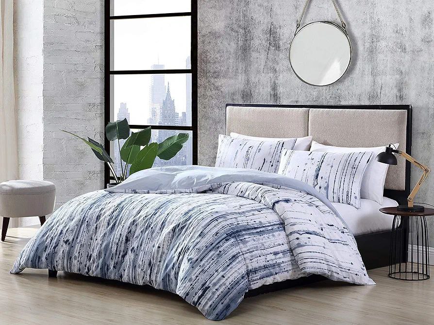 Industrial-style bedroom featuring a blue and white abstract duvet with matching pillows, set against a concrete accent wall and large window with city views. A modern duvet currently available for sale.