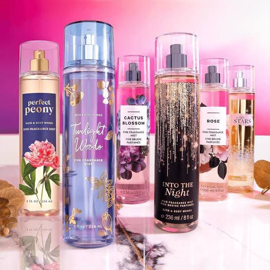 Bath and Body Works fragrance mist collection, featuring scents like Perfect Peony, Twilight Woods, Cactus Blossom, Into the Night, Rose, and In the Stars, displayed against a pink and purple backdrop.