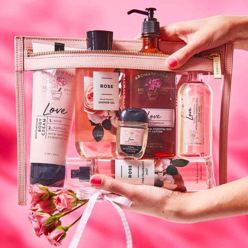 Bath and Body Works Rose and Aromatherapy Love collection in a pink zippered pouch, featuring body cream, shower gel, hand sanitizer, and essential oil lotion, held against a pink background.