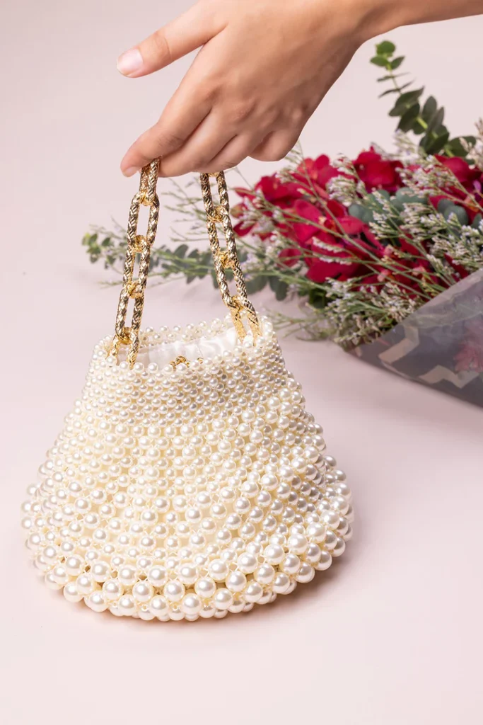 Chic pearl bucket bag with a gold chain handle, held by a hand, styled alongside red flowers for a feminine and elegant accessory.